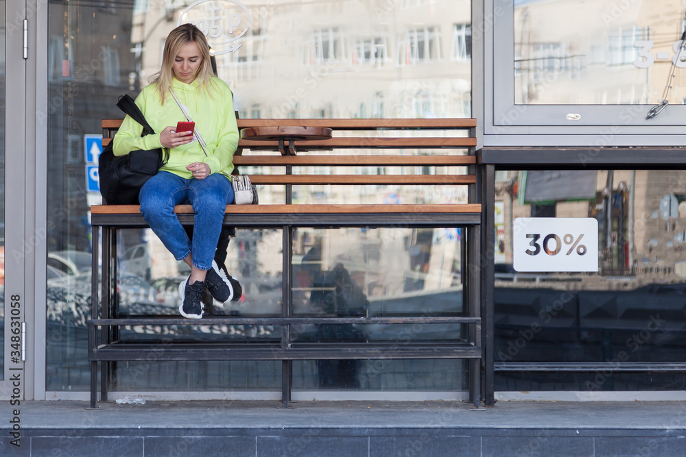 A blonde girl in a bright yellow sweatshirt sits on a wooden bench with a backpack near a coffee shop, holds a phone in her hands and waits for her order. The girl at the bus stop.