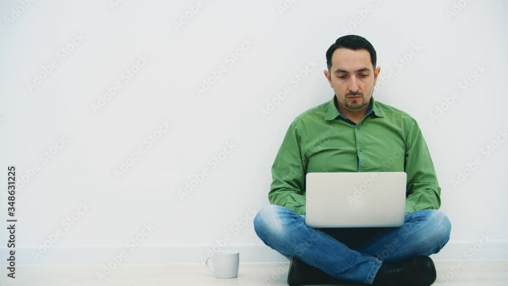 Isolated serious young man sitting on the floor in the lotos position with laptop and a white cup on his side, searching in the internet. Isolated serious young man sitting on the floor in the lotos position with laptop and a white cup on his side, searching in the internet.