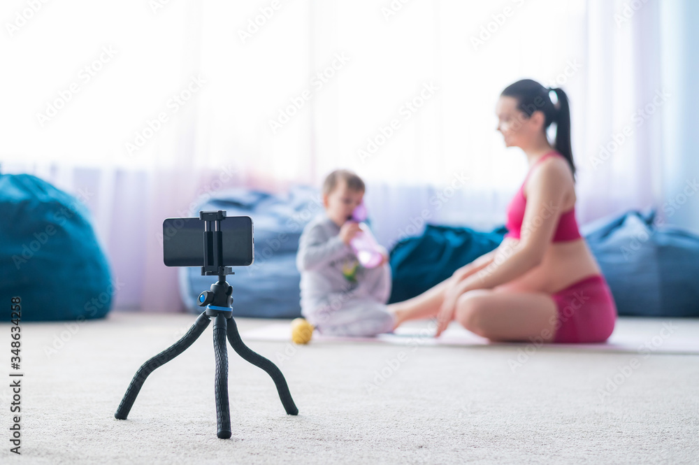 A pregnant woman does gymnastics with her son live. The expectant mother shoots a training video for her blog on the phone. Fitness with a baby.