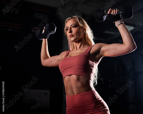 European woman blonde with a sports figure wearing gloves does biceps exercise with dumbbell. Fitness bikini model posing in the gym. Female bodybuilder.