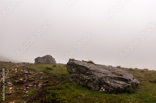 Large stones on a mountainside with fog in the background. Green grass on the ground. Rocky trail. Copy space. Horizontal.