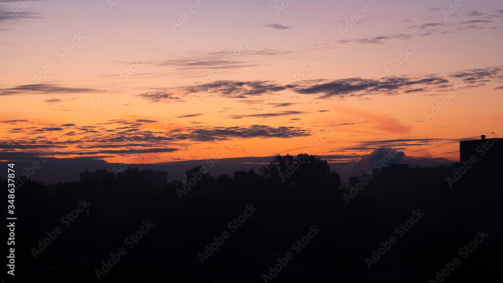 Sunrise over the dark city. Dark silhouettes of residential buildings and trees against a bright, orange sky with clouds.