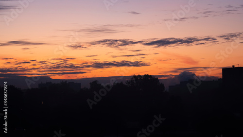 Sunrise over the dark city. Dark silhouettes of residential buildings and trees against a bright  orange sky with clouds.