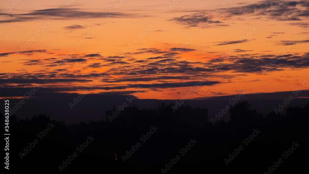 Fototapeta premium Sunrise over the dark city. Dark silhouettes of residential buildings and trees against a bright, orange sky with clouds.