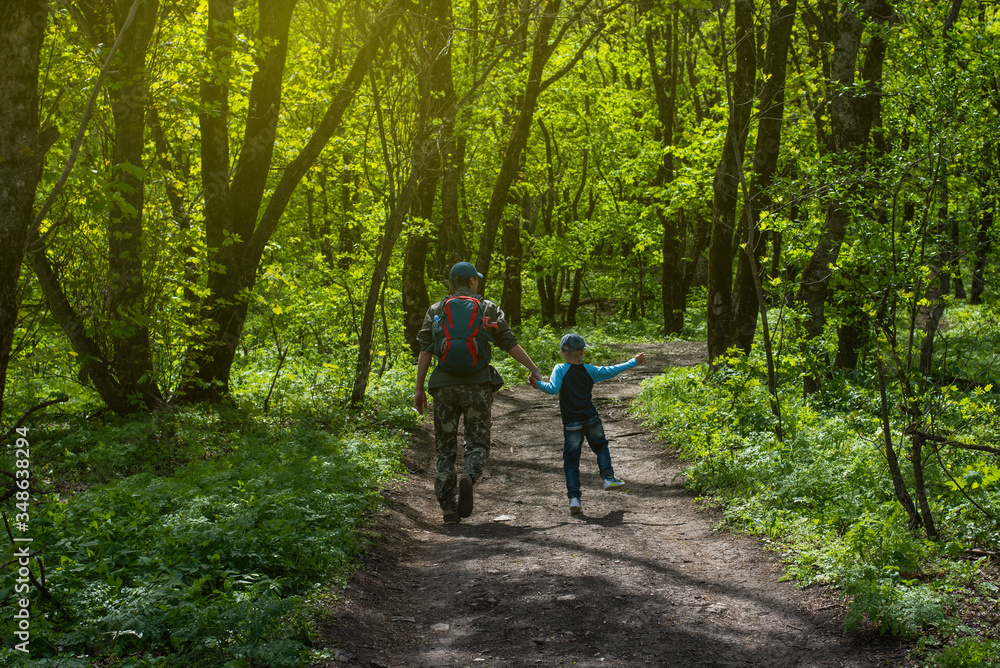 Forest green landscape in spring. Father and son walk along the path. View from the back, a man with a backpack, a boy runs in a jump. Family tourism. Card with copy space. Forest green landscape in spring. Father and son walk along the path. View from the back, a man with a backpack, a boy runs in a jump. Family tourism. Card with copy space.