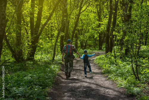 Forest green landscape in spring. Father and son walk along the path. View from the back, a man with a backpack, a boy runs in a jump. Family tourism. Card with copy space.