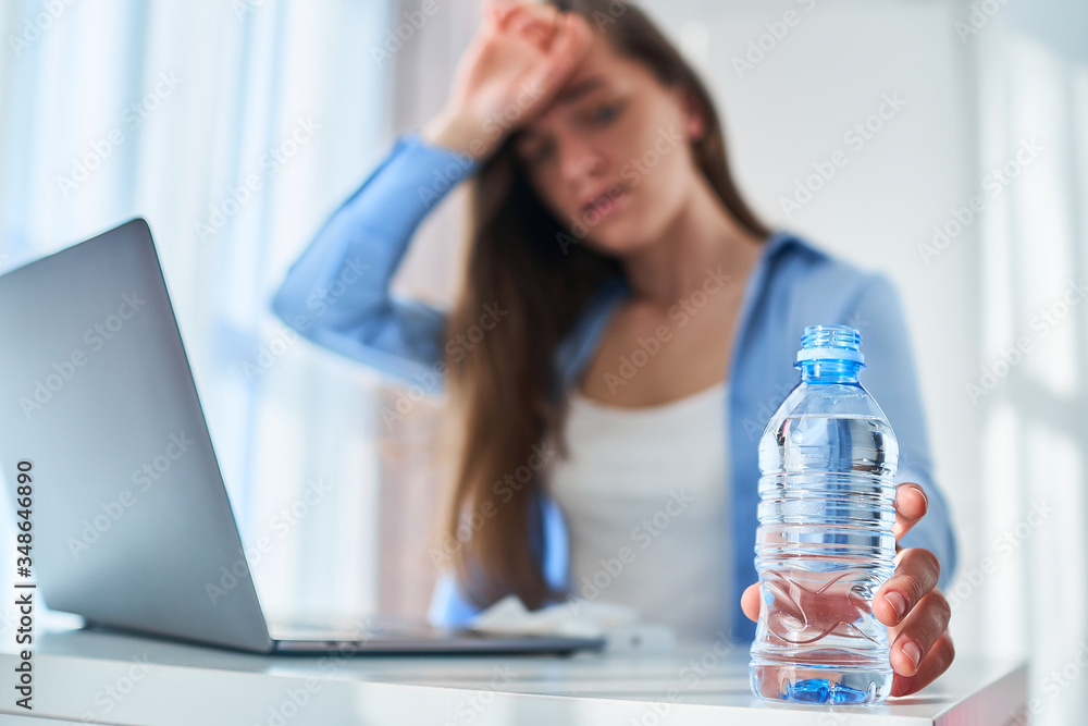 Upset tired working woman suffering from heat, thirst and hot weather cools down with cold water bottle during online work at computer at warm summer day - obrazy, fototapety, plakaty 