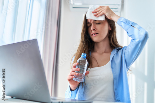 Sweating woman suffering from warm weather and thirst wipes forehead with a napkin and cools down with cold water bottle during online working at computer at hot summer day photo