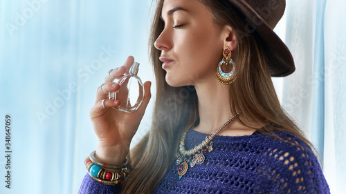 Stylish fashionable attractive brunette boho chic woman with closed eyes wearing jewelry and hat holds perfume bottle