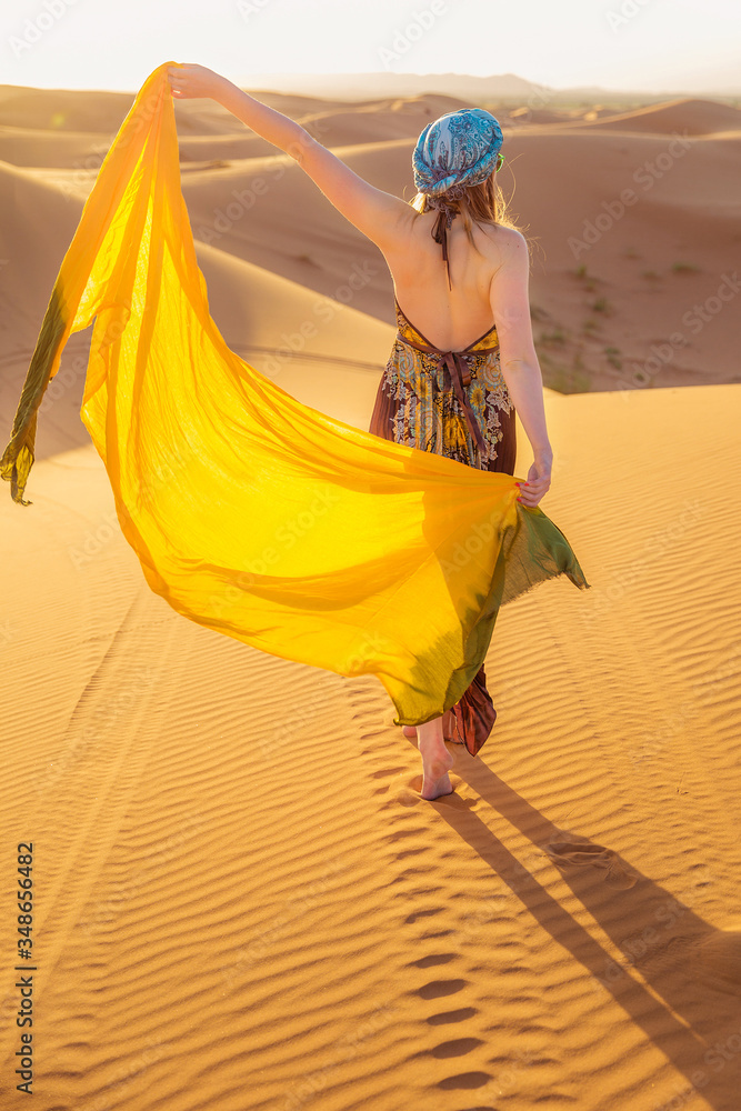 Obraz premium Beautiful young slim woman in a multi-colored dress with a yellow scarf, in a turban and sunglasses poses at dawn in the Sahara desert. Morocco Fashion.