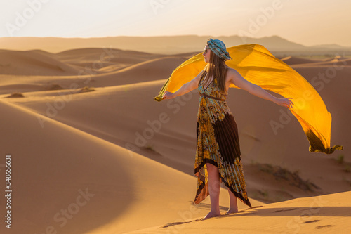 Beautiful young slim woman in a multi-colored dress with a yellow scarf  in a turban and sunglasses poses at dawn in the Sahara desert. Morocco Fashion.