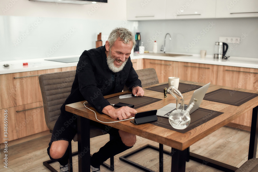 Time to chill. Cheerful middle-aged man holding a crutch, making a marijuana cigarette or joint, while sitting in the kitchen and using smartphone. Bong, lighter, cup and laptop on the table Time to chill. Cheerful middle-aged man holding a crutch, making a marijuana cigarette or joint, while sitting in the kitchen and using smartphone. Bong, lighter, cup and laptop on the table