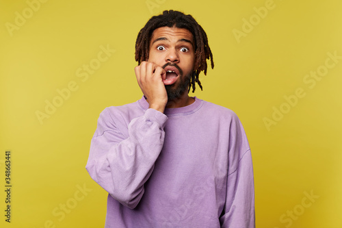 Afraid young attractive brown haired bearded man with dark skin looking at camera with wide eyes opened and holding scaredly hand on his face, isolated over yellow background