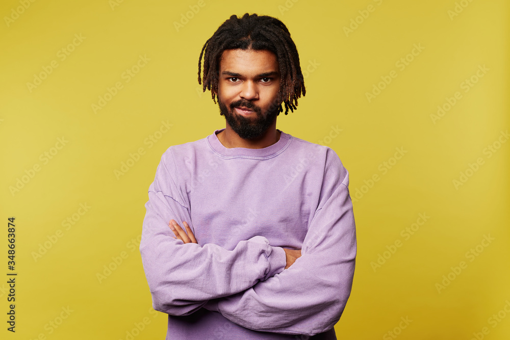Offended young pretty dark skinned brunette male in purple sweatshirt crossing arms on chest and twisting mouth while looking at camera, isolated over yellow background