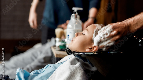 Visiting barbershop. Side view of a cute boy leaning on the sink while male barber washing his hair before cutting. Child in hairdressing salon