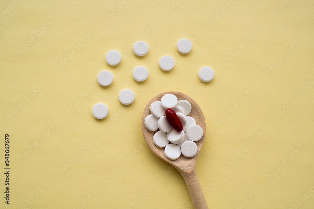 round white tablets with one red capsule on a wooden spoon on a yellow background, tablets are scattered in the center from bottom to top, isolated. round white tablets with one red capsule on a wooden spoon on a yellow background, tablets are scattered in the center from bottom to top, isolated.