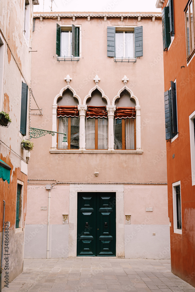 Facade of a three-story peach house in Venice, Italy. Massive green wooden front door, three classic Venetian windows with a sharp top, columns and rag blinds.