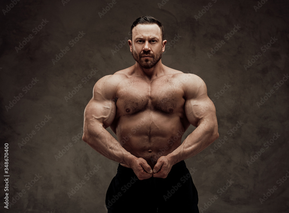 Shirtless adult male bodybuilder doing a muscle showing with a serious face in a studio
