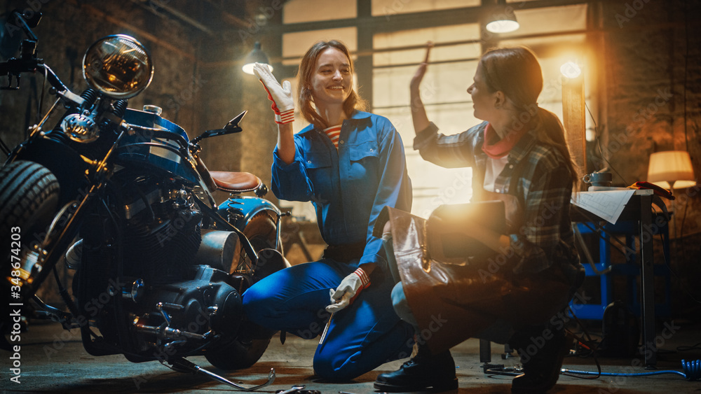 Two Young Beautiful Female are Discussing The Work Done on a Custom Bobber Motorcycle. Talented Girls Use a Tablet Computer. They are Happy and Doing a High Five. Creative Authentic Workshop Garage.