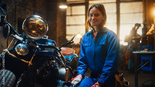 Young Beautiful Female Mechanic is Working on a Custom Bobber Motorcycle. Talented Girl Wearing a Blue Jumpsuit. She Smiles at the Camera. Creative Authentic Workshop Garage.