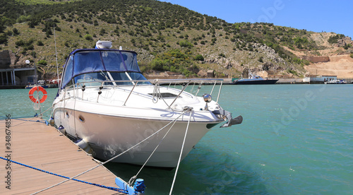 A white motor boat with two decks is moored to the pier. The upper tier is covered with a blue awning. The boat is lowered to the sea  behind the boat is a mountainous landscape. Removed from the pier