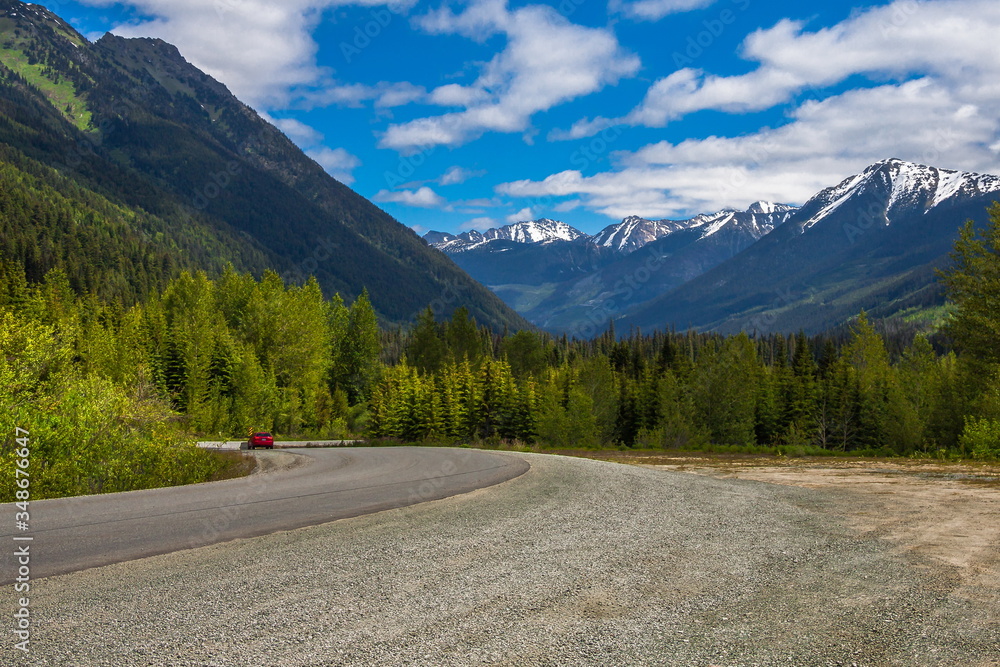 Fototapeta premium Highway from sea to sky. A car drives down a mountain road to the Lillooet Town, deciduous forest on the sides of the road a mountain range on the horizon against a blue cloudy sky