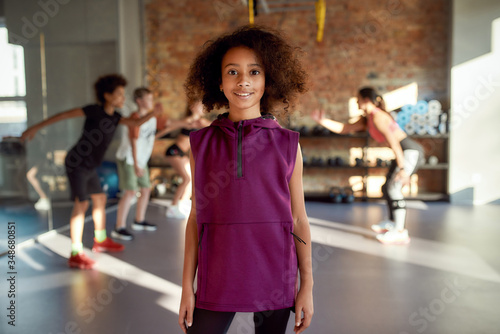 Little sportswoman. Portrait of a girl smiling at camera before warming up, exercising together with other kids and trainer in gym. Sport, healthy lifestyle, active childhood concept