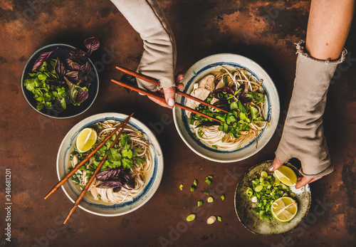 Asian cuisine lunch. Flat-lay of Vietnamese rice noodle chicken soup Pho Ga with cilantro, soy sprouts, greens and womans hands with chopsticks and fresh lime over dark rusty background, top view