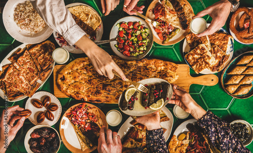 Muslim Ramadan iftar family dinner. Flat-lay of people feasting over table with Middle East food. Dates, dolma, kebab, flatbread, pide, borek, sweet, salad, top view. Ramazan fasting Turkish cuisine