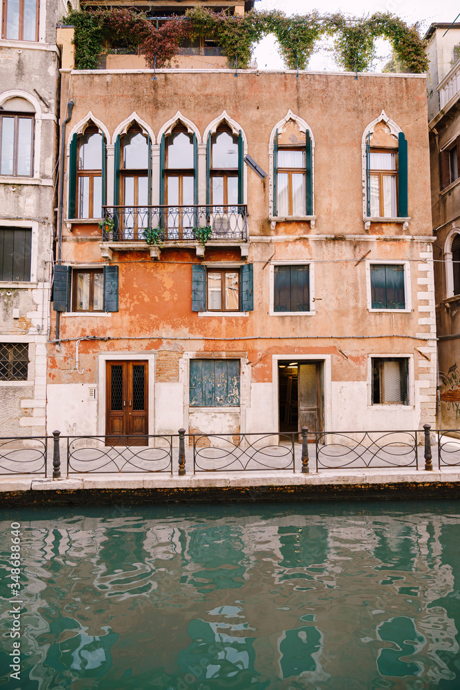 Close-ups of building facades in Venice, Italy. An ancient stone three-storey house on the banks of Venetian Canal. Metal wrought-iron fence, near sea water. Classic Venetian windows, with sharp tops