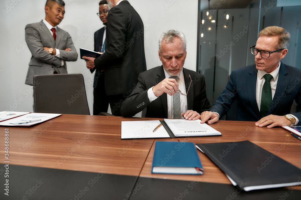 I have to think. Confident mature businessman holding eyeglasses and reading a contract while having a meeting with his business partners in the modern office I have to think. Confident mature businessman holding eyeglasses and reading a contract while having a meeting with his business partners in the modern office