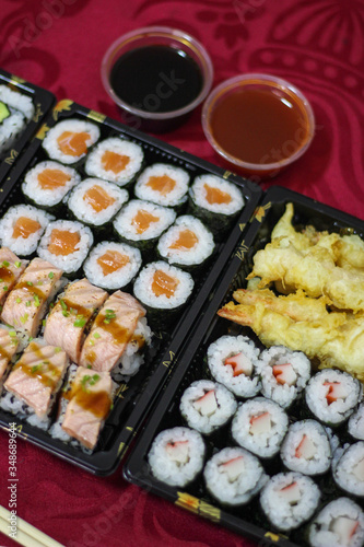 Sushi take-away delivery boxes with salmon Hosomaki, Californias, URAMAKI, breaded shrimp, tempura shrimp, bowl of bittersweet, sweet and sour sauce, chopsticks on a red tablecloth vertical