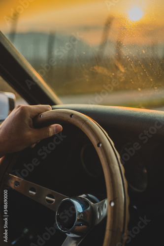 Close up od an unrecognisable man holding a wooden steering wheel of a vintage muscle car driving into the sceninc sunset. Idyllic rural landscape is visible. photo