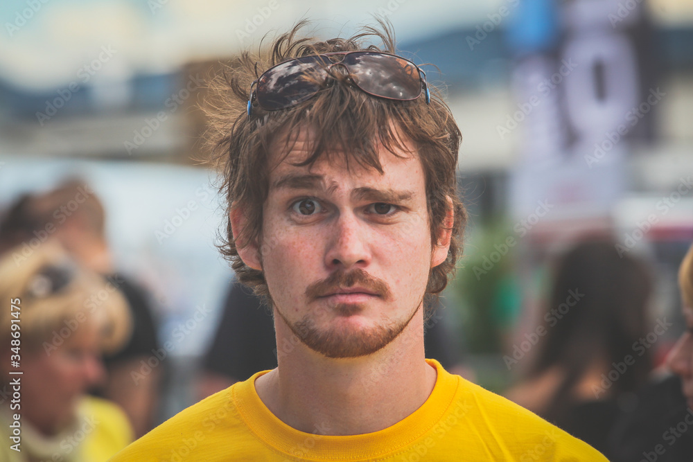 Confused young caucasian man with moustache, a yellow tshirt and sunglasses in his hair in a public space, looking towards the camera. Confused young caucasian man with moustache, a yellow tshirt and sunglasses in his hair in a public space, looking towards the camera.
