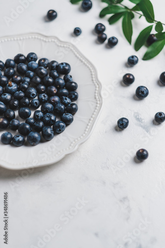 Fresh juicy blueberries in a white plate with green leaves on a white background. 