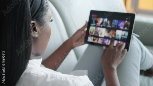 African American woman lying down on sofa in living room at home searching for film or TV series to watch for leisure. Black lady choosing movie on internet streaming service. Over the shoulders view