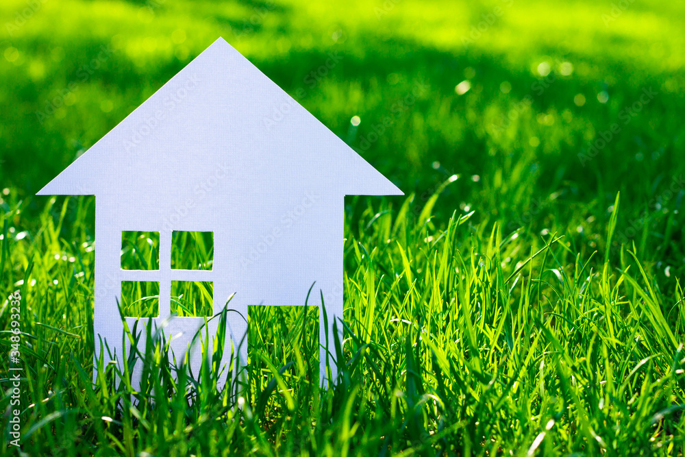 White silhouette of a toy house cut out of cardboard on a background of grass. A small house made of white paper. Copy space. White silhouette of a toy house cut out of cardboard on a background of grass. A small house made of white paper. Copy space.