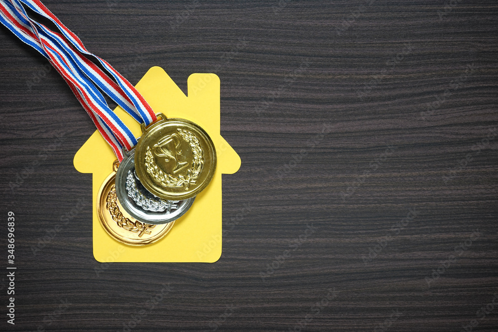 Set of gold, silver and bronze medals with house replica made from yellow cardboard on top of dark brown wooden pattern background Set of gold, silver and bronze medals with house replica made from yellow cardboard on top of dark brown wooden pattern background