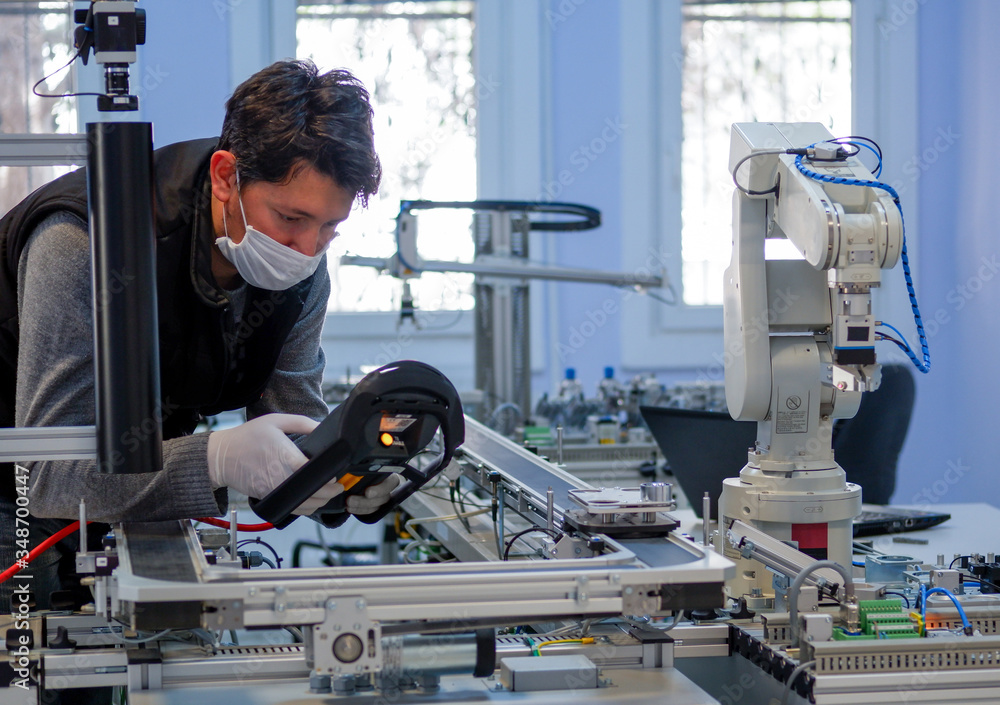 Man with surgical mask on face against COVID-19 ( Coronavirusis ) programming robot arm with control panel which is integrated on smart factory production line. industry 4.0 automation line. Man with surgical mask on face against COVID-19 ( Coronavirusis ) programming robot arm with control panel which is integrated on smart factory production line. industry 4.0 automation line.