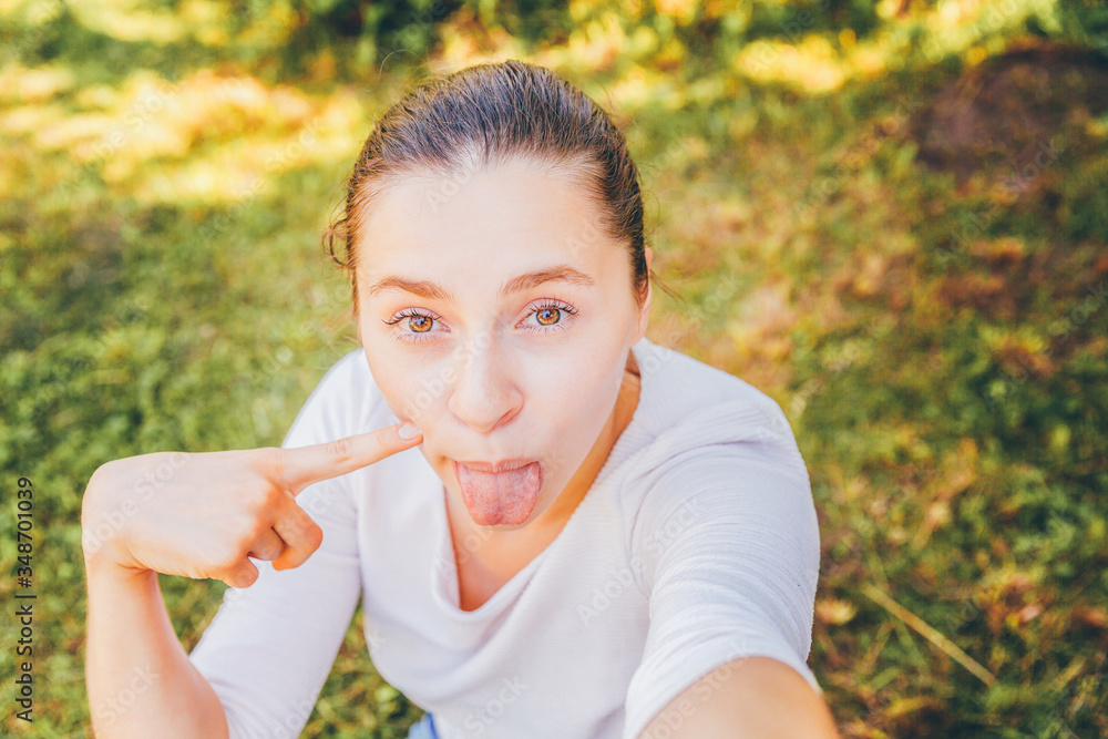 Fototapeta premium Young girl take selfie from hands with phone showing tongue and funny face sitting on park or garden background. Portrait of young attractive woman making selfie photo on smartphone in summer day.