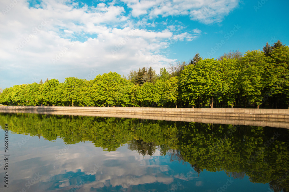 A lake in the city park early in the morning. Quiet and peaceful, in harmony, no people. Fresh green trees and grass, blue sky and clouds reflecting in the water. Warm sun light. Minsk Belarus A lake in the city park early in the morning. Quiet and peaceful, in harmony, no people. Fresh green trees and grass, blue sky and clouds reflecting in the water. Warm sun light. Minsk Belarus
