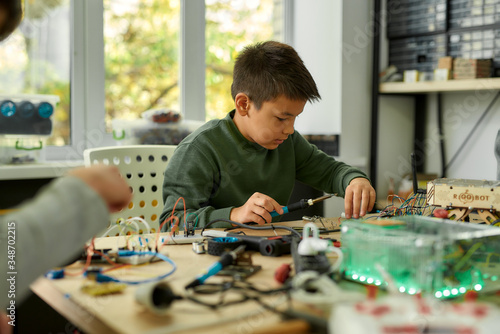 Nothing great ever come easy. Asian boy using soldering iron to join chips and wires while making his own vehicle at a stem robotics class. Inventions and creativity for children.