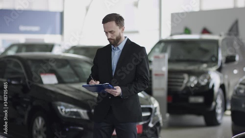 Auto dealership manager is counting cars. Young salesman writing on his clipboard. Handsome bearded man in shirt and black suit is observing cars