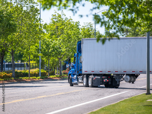 Blue day cab big rig semi truck with roof spoiler transporting cargo in dry van semi trailer turning on the green city street road