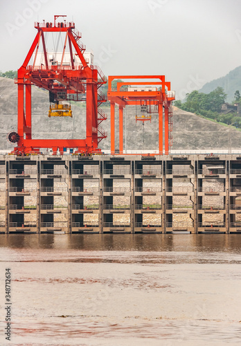 Huangqikou, Chongqing, China - May 8, 2010: Yangtze River. Closeup of red cranes on multi-level port quay behind brown water. hill with green foliage in back under gray sky. photo