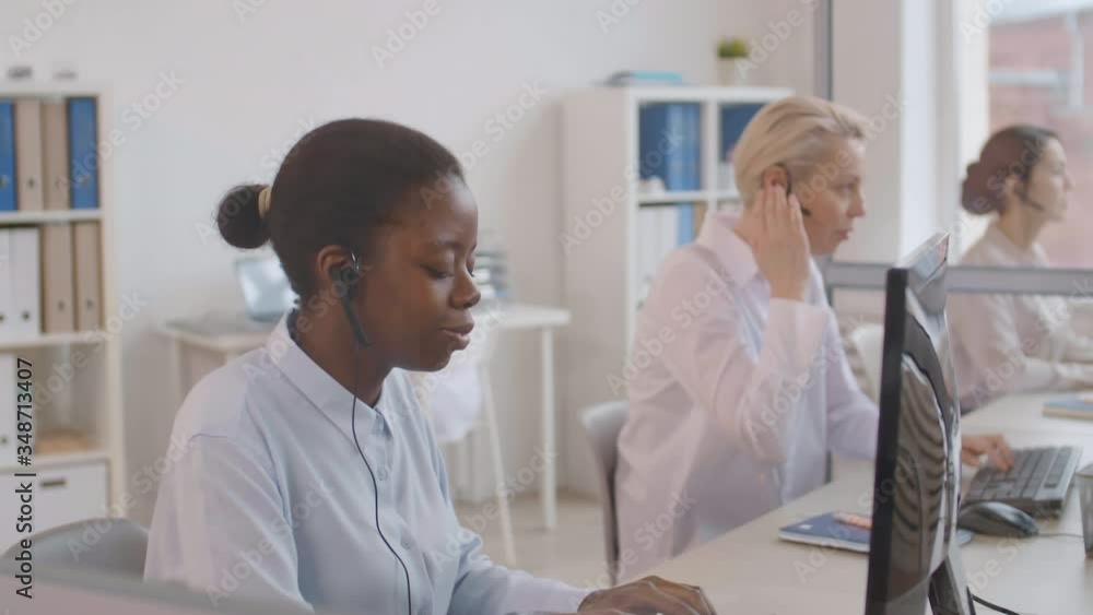 Medium shot of three diverse women wearing white shirts and headsets sitting at computer monitors in the office and consulting costumers by telephone Medium shot of three diverse women wearing white shirts and headsets sitting at computer monitors in the office and consulting costumers by telephone