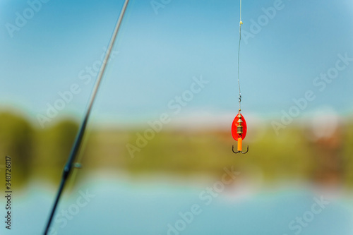 A bright spinner with a triple hook for fishing hangs on a fishing line above the water. Selective focus.