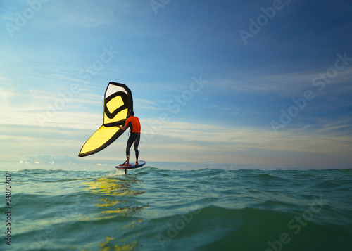 A man wingfoils in the sea on a summer's day, using a hand held inflatable wing and riding a hydrofoil surfboard.  Green sea with cloudy blue sky background photo