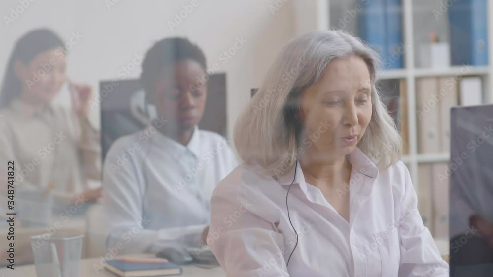 Medium shot of elderly Asian woman wearing headset sitting at PC, talking to client then looking at camera and smiling while her colleagues working on background Medium shot of elderly Asian woman wearing headset sitting at PC, talking to client then looking at camera and smiling while her colleagues working on background