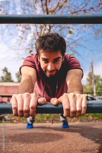 Handsome young man portrait smiling and training with focus on hands grabbed from bar on a calisthenics park in a sunny day with blue sky